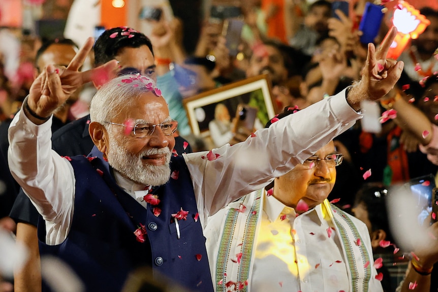 Narendra Modi in a white shirt and blue vest holds his arms in the air making peace signs with his fingers at a rally