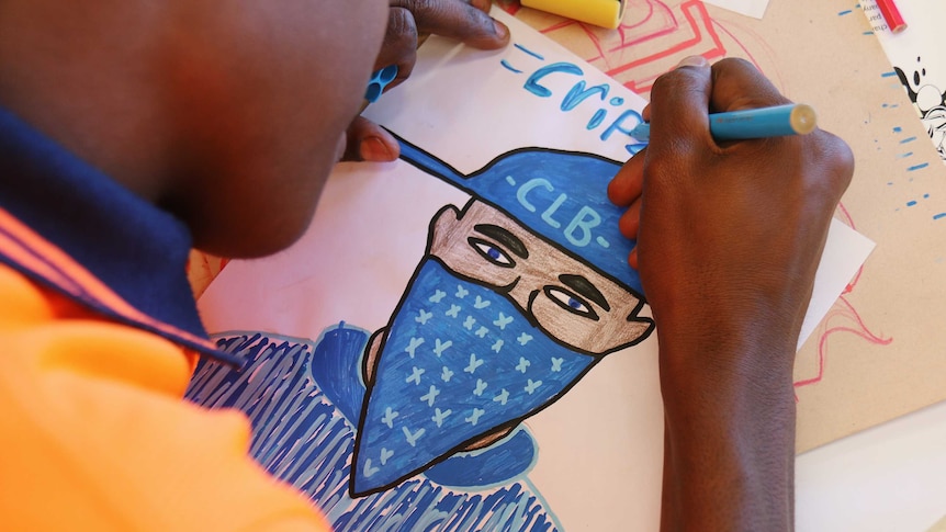 A young man draws a figure in a blue bandana with the word 'crips' above it.