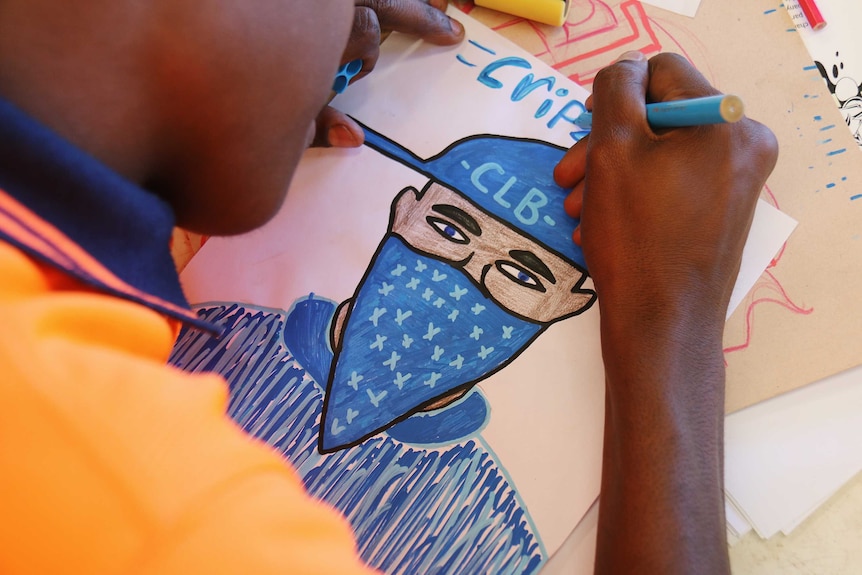 A young man draws a figure in a blue bandana with the word 'crips' above it.