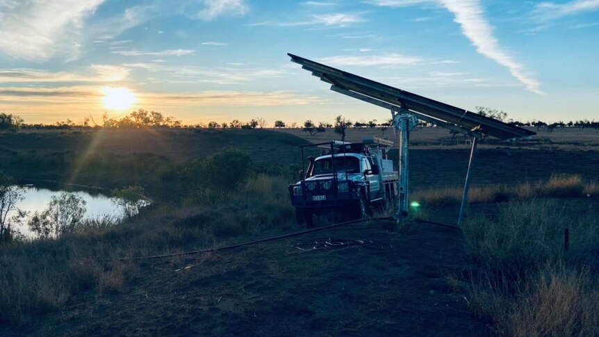 Car parked under solar panels next to a dam with the sun setting in the background. 