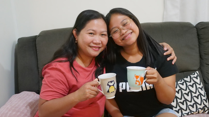 A mother and daughter sitting on a couch holding mugs, the mother's arm is around the daughter's shoulders