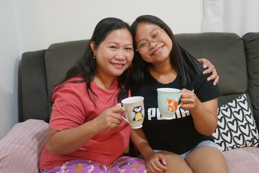 A mother and daughter sitting on a couch holding mugs, the mother's arm is around the daughter's shoulders