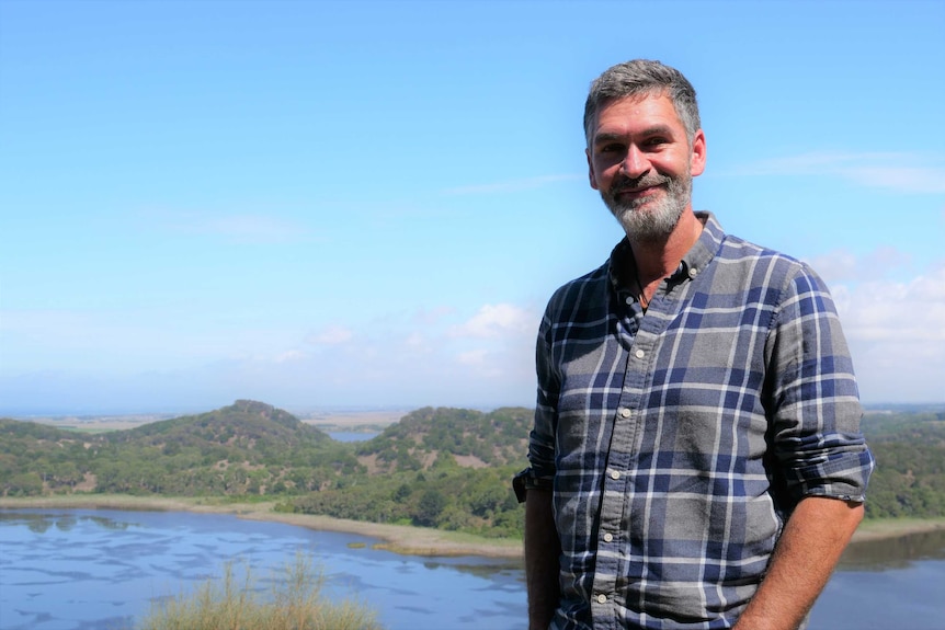 A middle-aged man stands on a hill with a lake behind him.