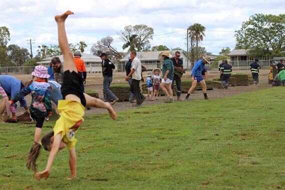 A young student does cartwheel on new turf with volunteers laying turf in background.