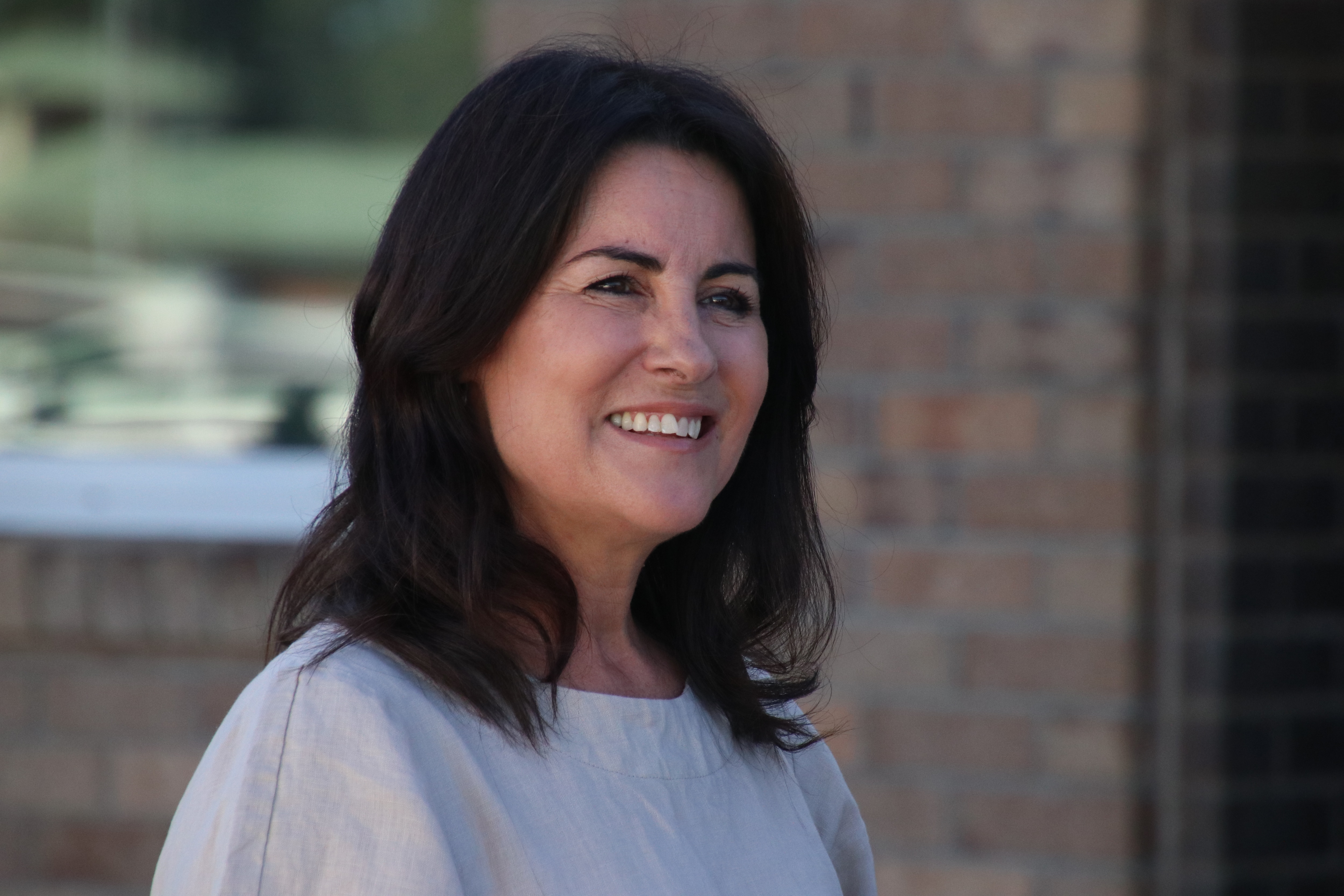 Uma mulher de cabelos castanhos está parada em frente a uma parede de tijolos marrons, sorrindo.