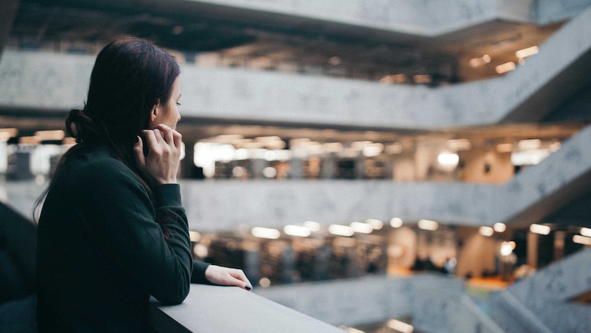 Woman leans on balcony with head on hand, blurred background with people