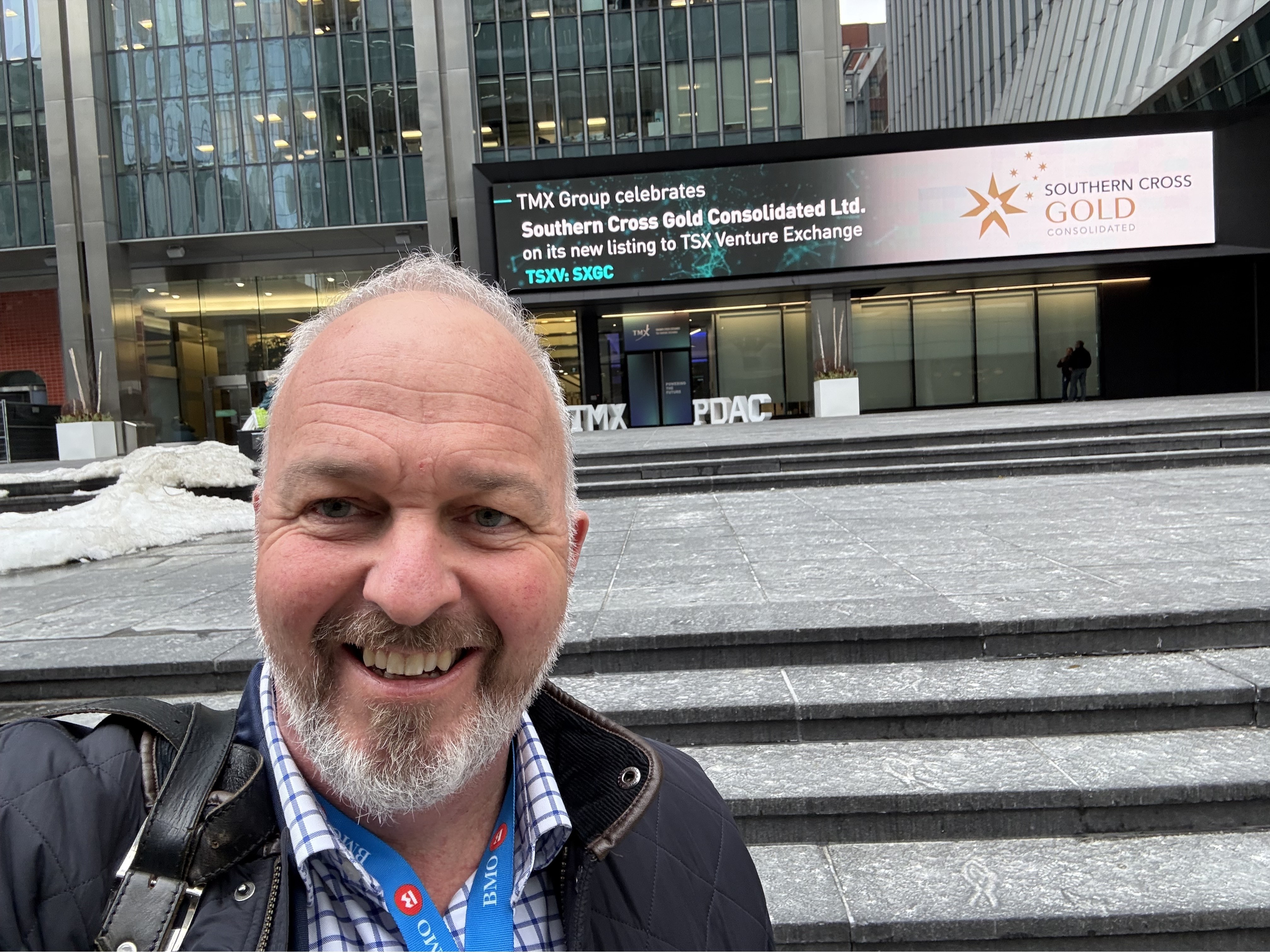 Un hombre con barba sonríe en una foto selfie frente a un cartel publicitario.