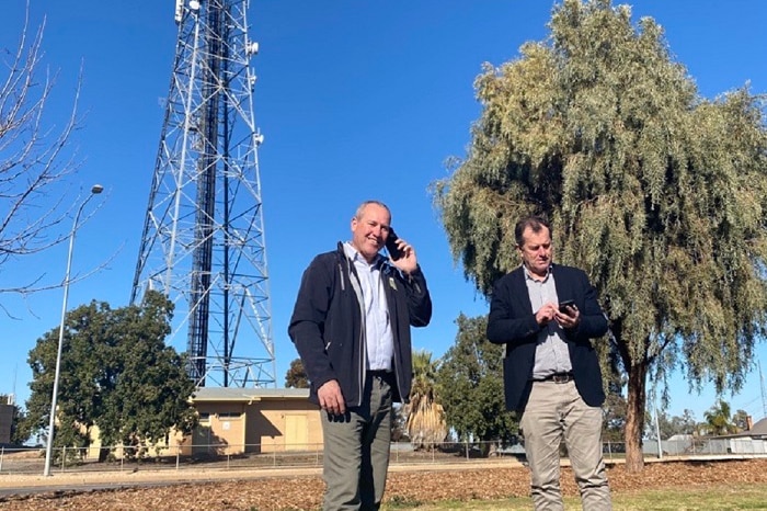 Two men stand next to each other on their phones.