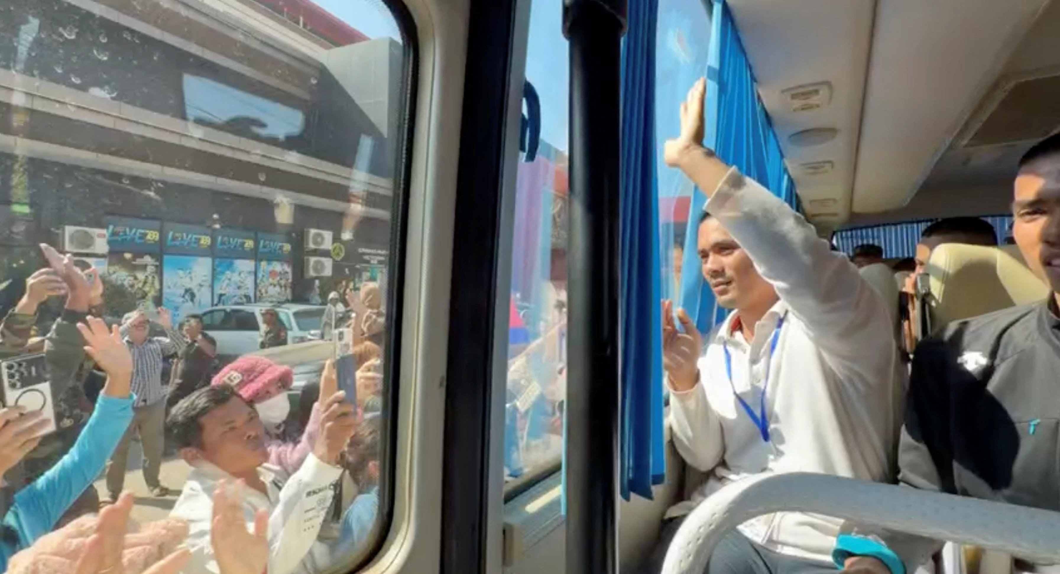 Un hombre con camisa blanca mira por la ventana de un autobús a la gente que lo filma en la calle.