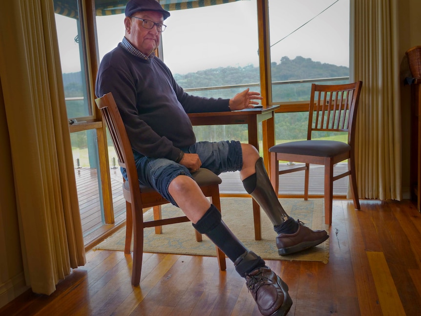 An older man sitting at a table by a window. He has poly-carbon leg splints