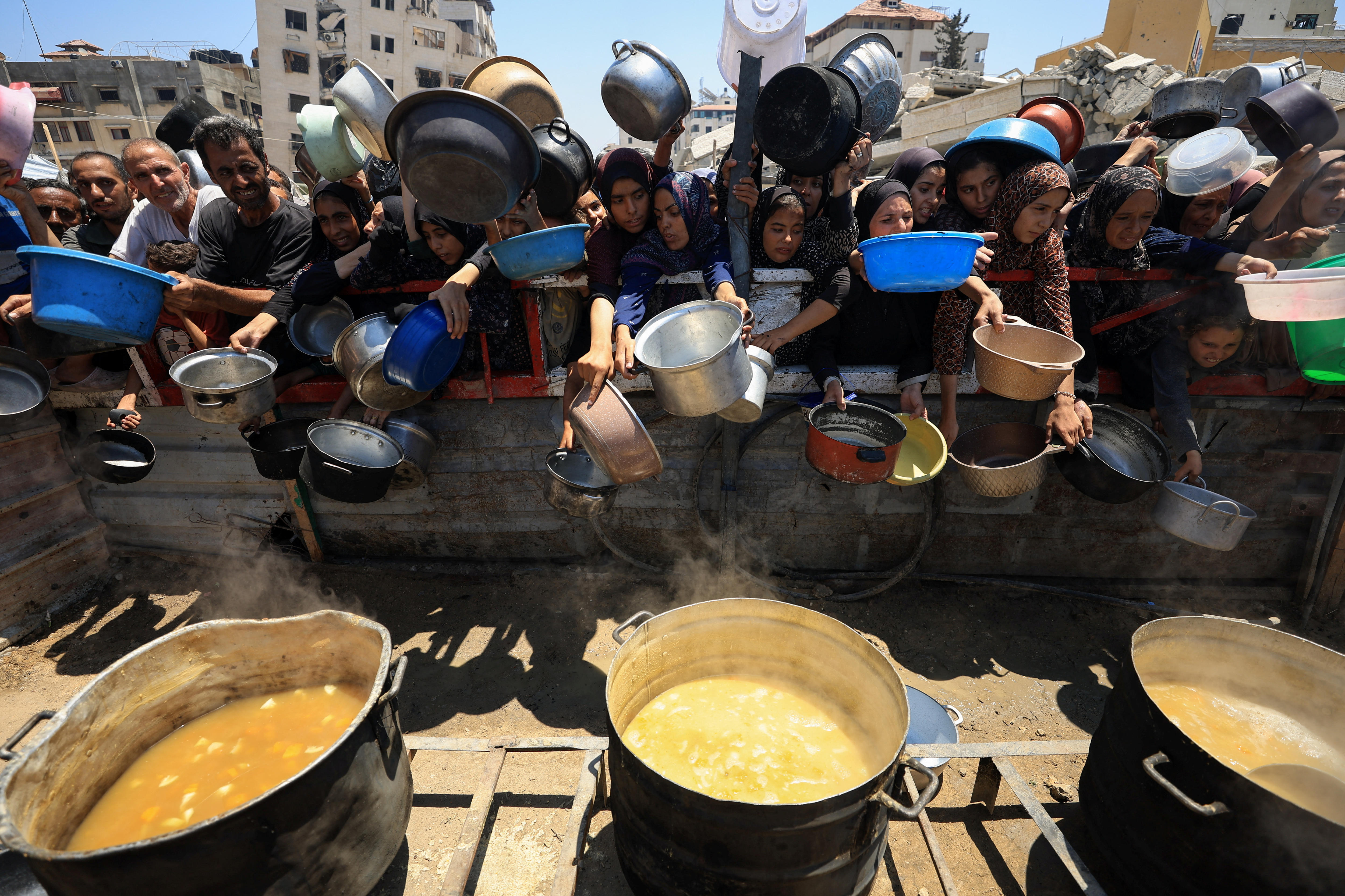 A crowd of Palestinians standing behind a barricade with three large pots of food in front of them.