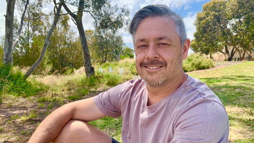 Simon Corcoran smiles as he sits in grassy parkland in a t-shirt.