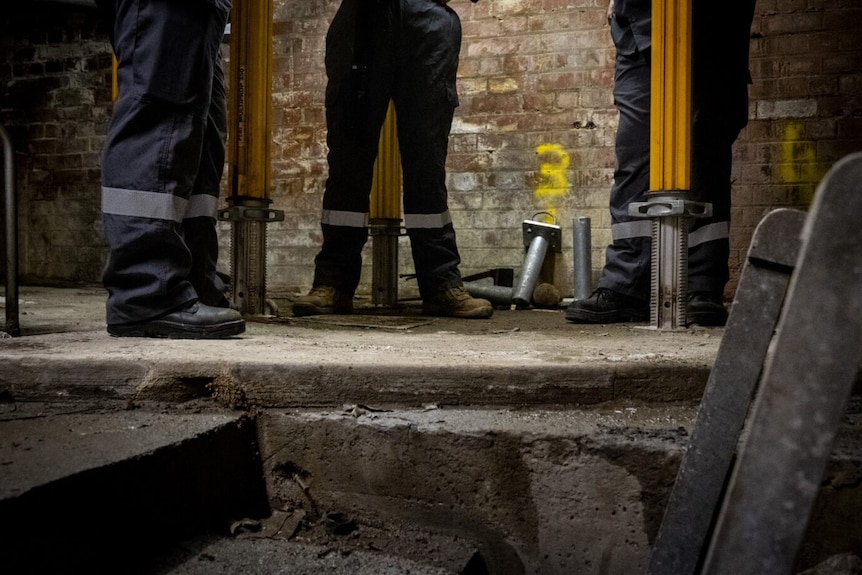 Three workers' legs are shown, standing in a tunnel underneath Melbourne.