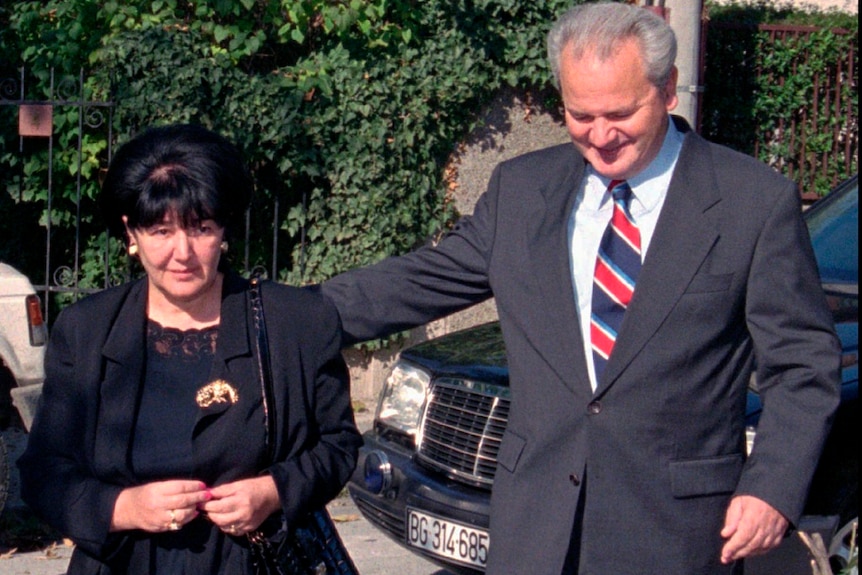 A middle aged man and woman wearing formal outfits walk from parked cars.