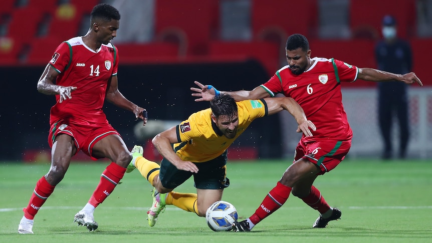 A soccer player in a yellow top falls forward as two defenders in red tops take a white ball from just in front of his legs.