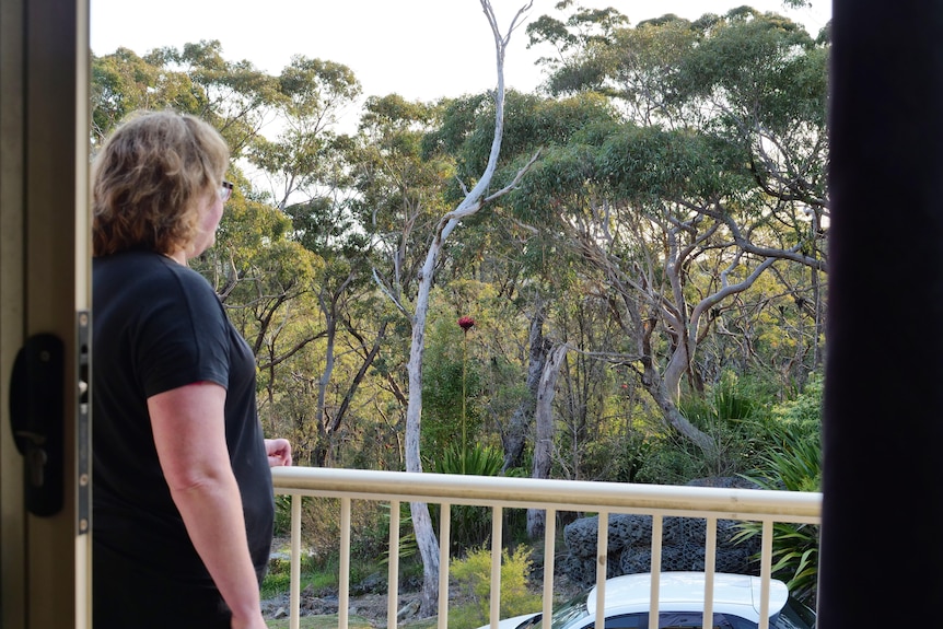 A woman standing on a balcony looks away at nearby bushland.