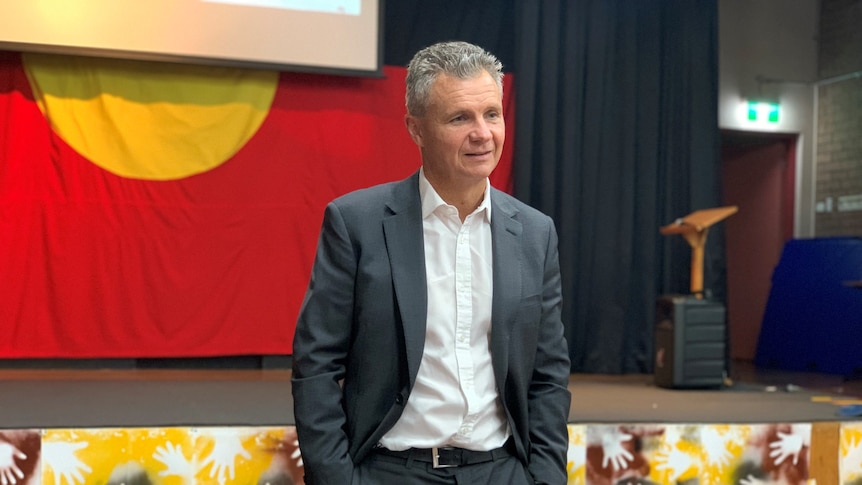 A man in a suit in front of an Aboriginal flag