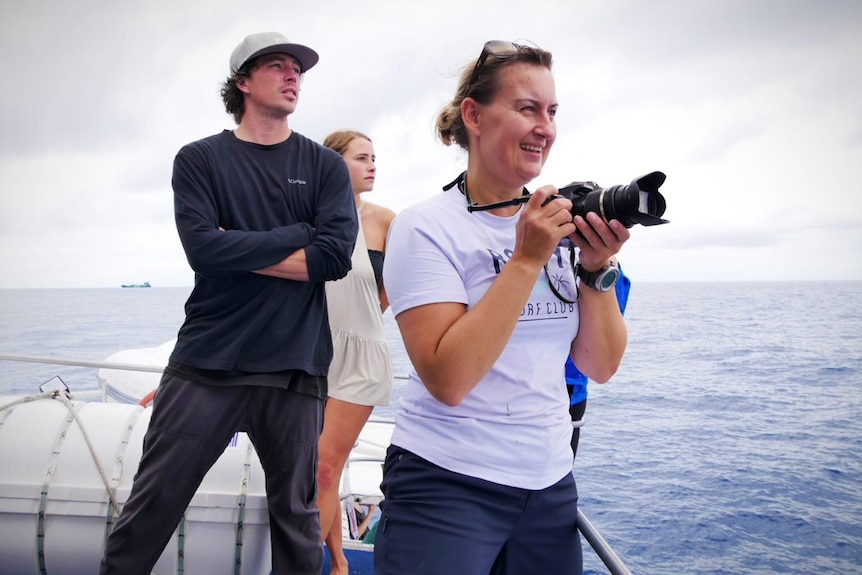 A smiling woman, holding a camera on a boat, looks to sea in front of a young man and woman.