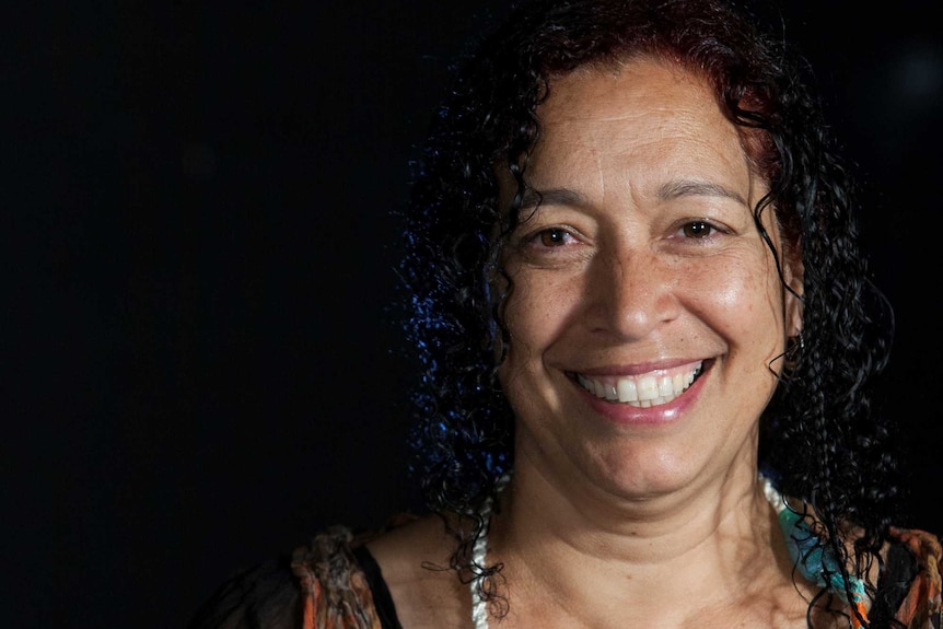 Rachael Maza smiles at the camera in front of a black backdrop.