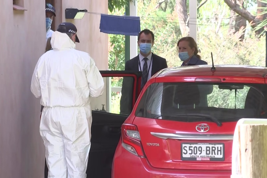 One police officer in a forensics suit and others wearing face masks next to a red car and a house