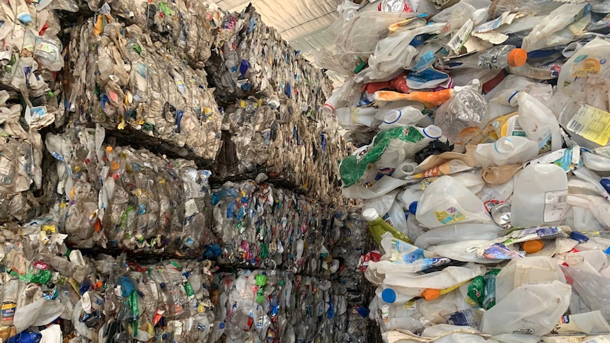 On the left of the screen recycling is in  bundles, stacked to the ceiling. On the right loose plastic contains are stacked.