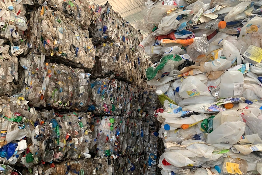 On the left of the screen recycling is in  bundles, stacked to the ceiling. On the right loose plastic contains are stacked.