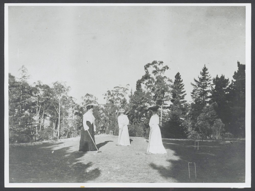 fotografia em preto e branco de mulheres em vestidos brancos na grama, com aros, bolas e marretas cercadas por árvores