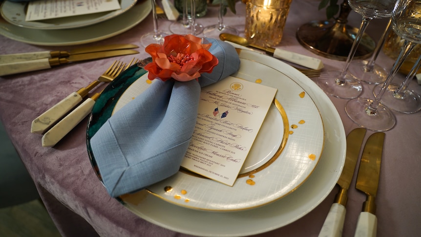 A plate with an ornate menu sits on a table with floral decorations and gold cutlery