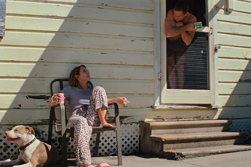 A 40-something man leans out of a back door to talk to a brown-haired woman who is seated outside with a dog beside her