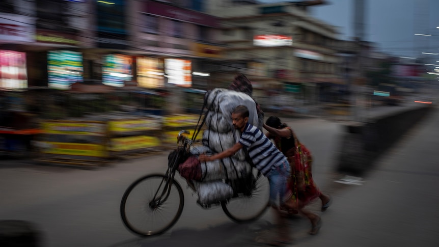 An Indian couple push a bicycle loaded with heavy white bags through a town street at night