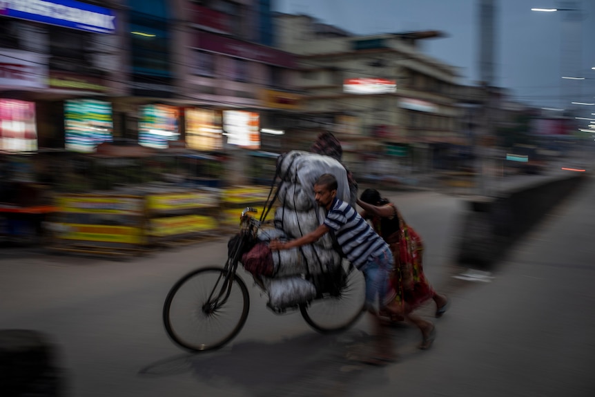 An Indian couple push a bicycle loaded with heavy white bags through a town street at night