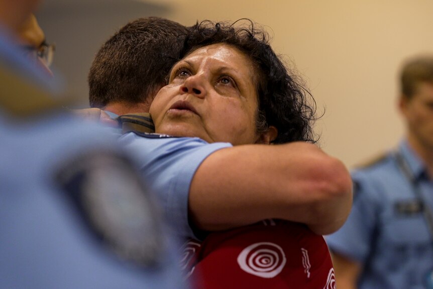 A woman looks up to the ceiling with an emotional expression while hugging a police recruit