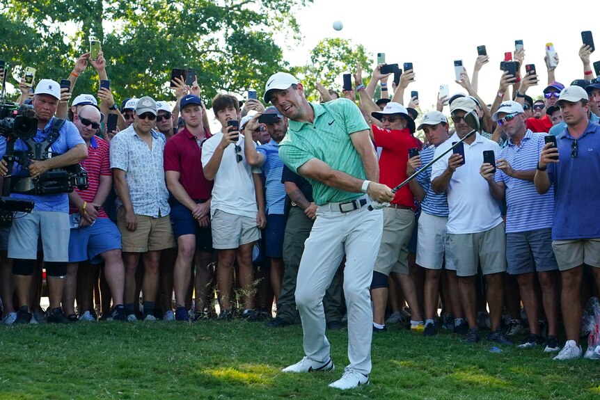 Golfer Rory McIlroy chips the ball in the air as a big group of spectators watch from close behind. 