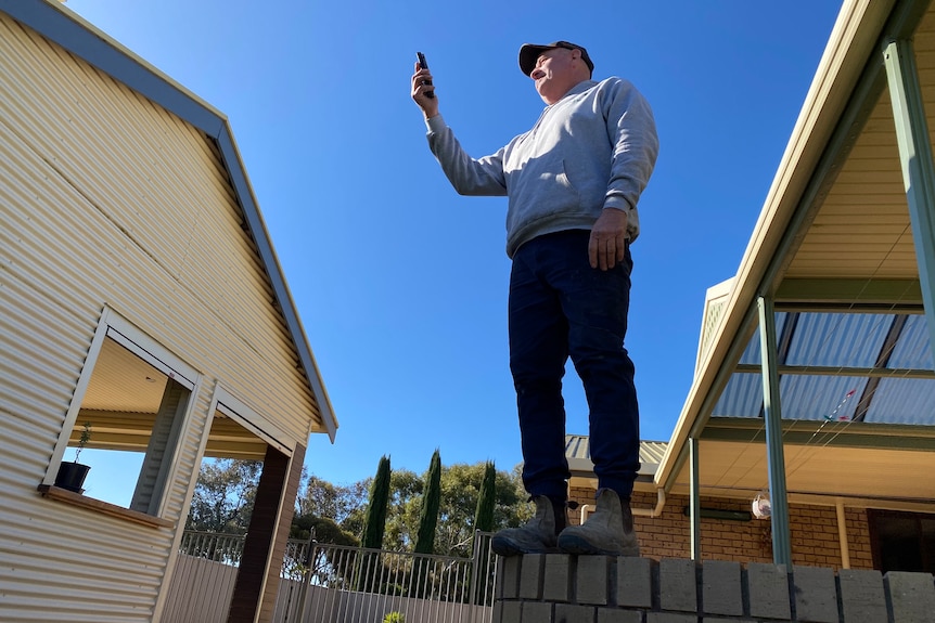 A man holds a phone up in air, he wears grey jumper, work pant and boots. He stands on a stone fence facing a roof