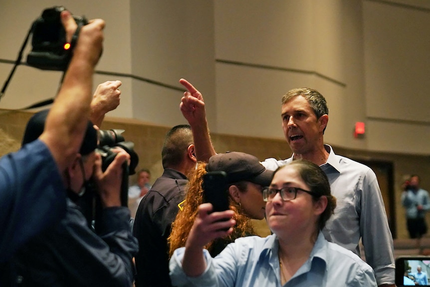 Beto O'Rourke, in a pale blue shirt, points his finger with a stern, angry expression on his face. People take photos