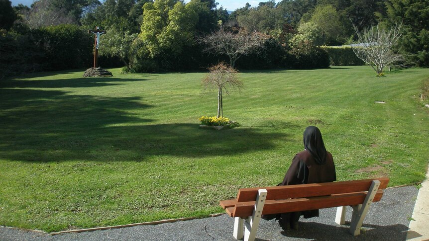 A nun sits outside on a seat looking into a garden.