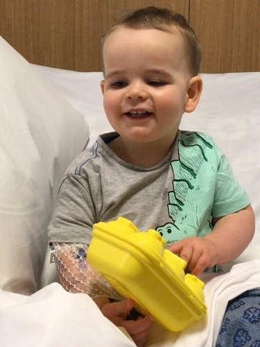 Rohan, a toddler, grins, sitting in a hospital bed with some bandages on.