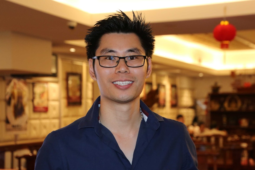 Quan Chen stands in his restaurant, a red lantern and diners around a table can be seen in the background.