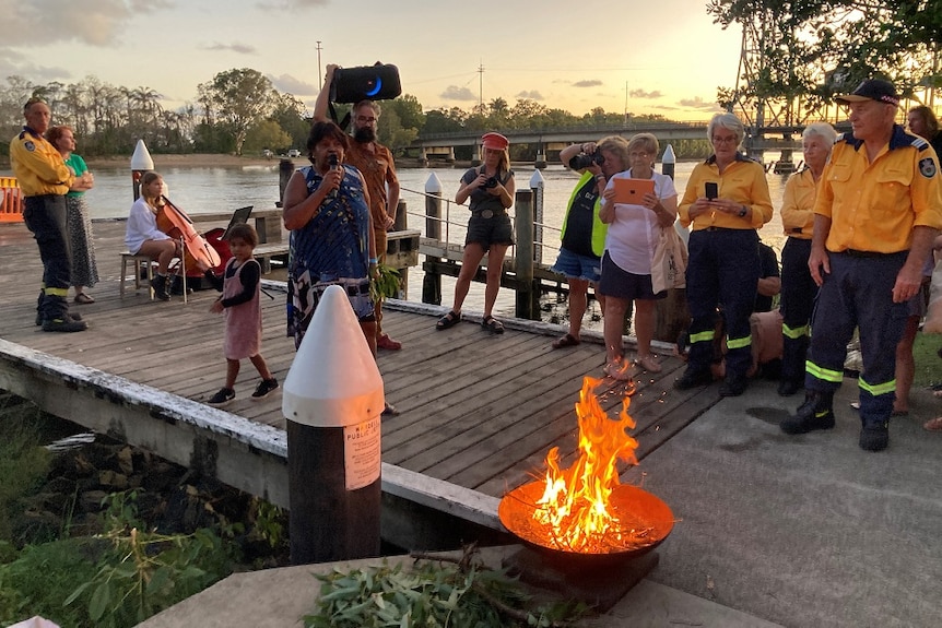 People stand on a wooden walkway near a river with an open fire nearby. 