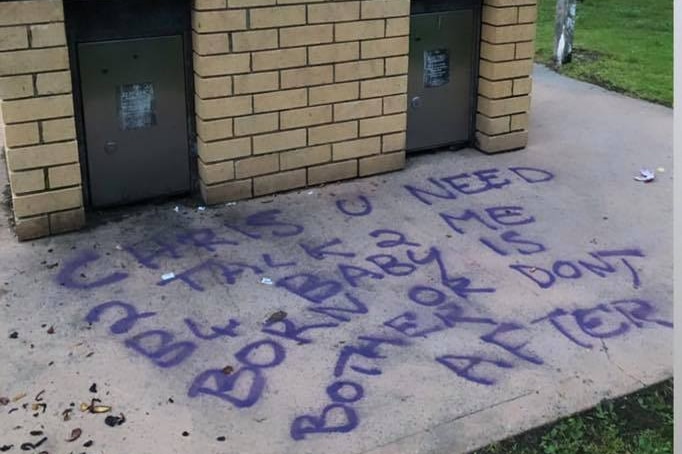 Graffiti sprayed onto the ground near a BBQ in a park reads 'Chris u need 2 talk 2 me b4 baby is born or dont bother after'.