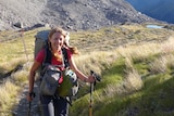 A smiling woman in hiking clothes and holding walking sticks stands before large mountains stretching back into the distance.