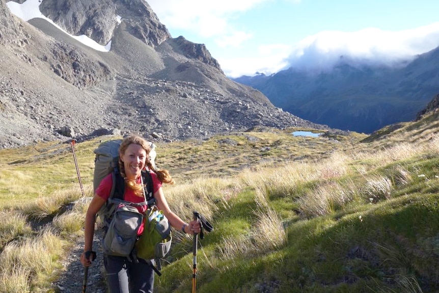 A smiling woman in hiking clothes and holding walking sticks stands before large mountains stretching back into the distance.