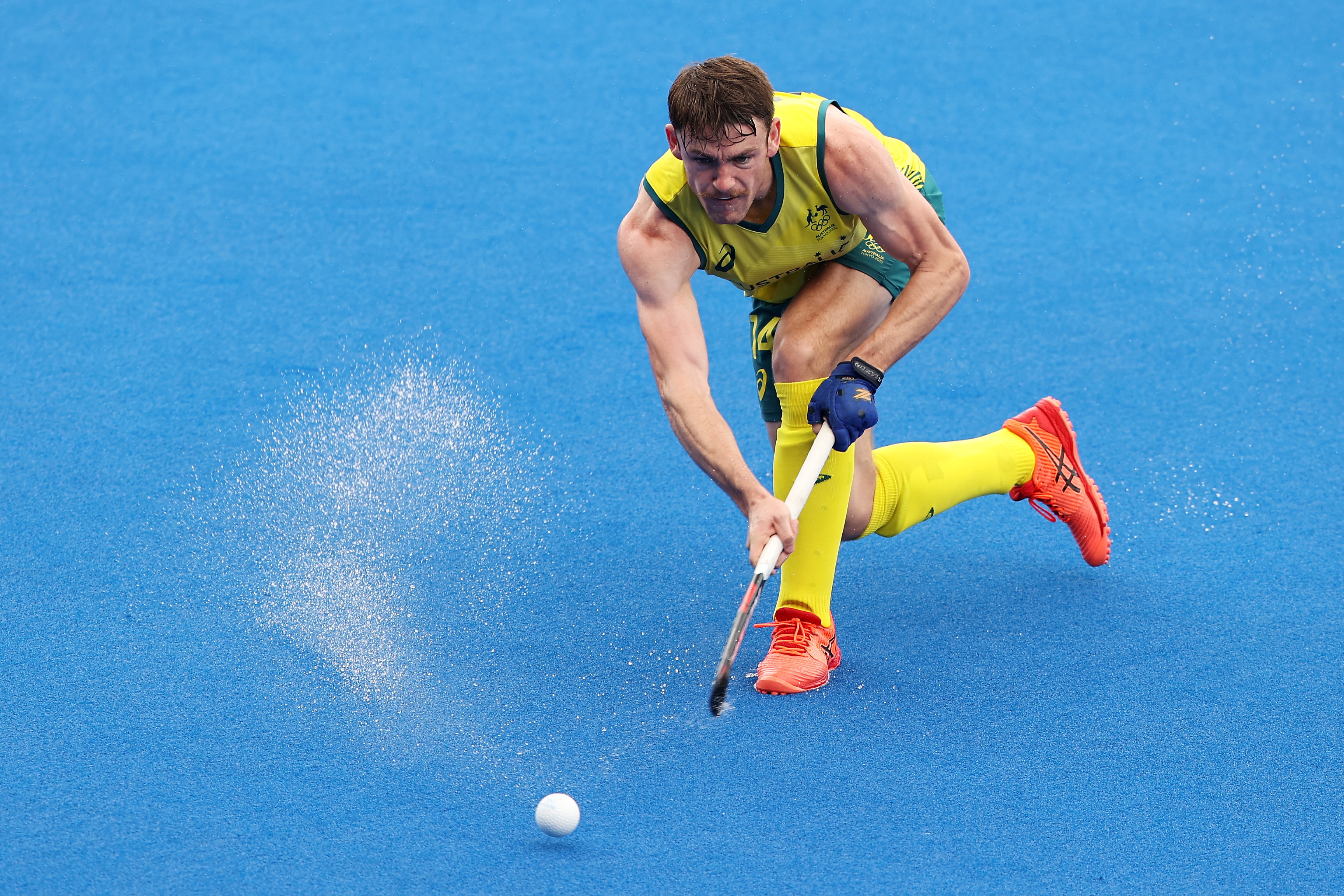 Man wearing yellow socks, green and yellow shorts and singlet, orange sneaker, chases a ball with a stick