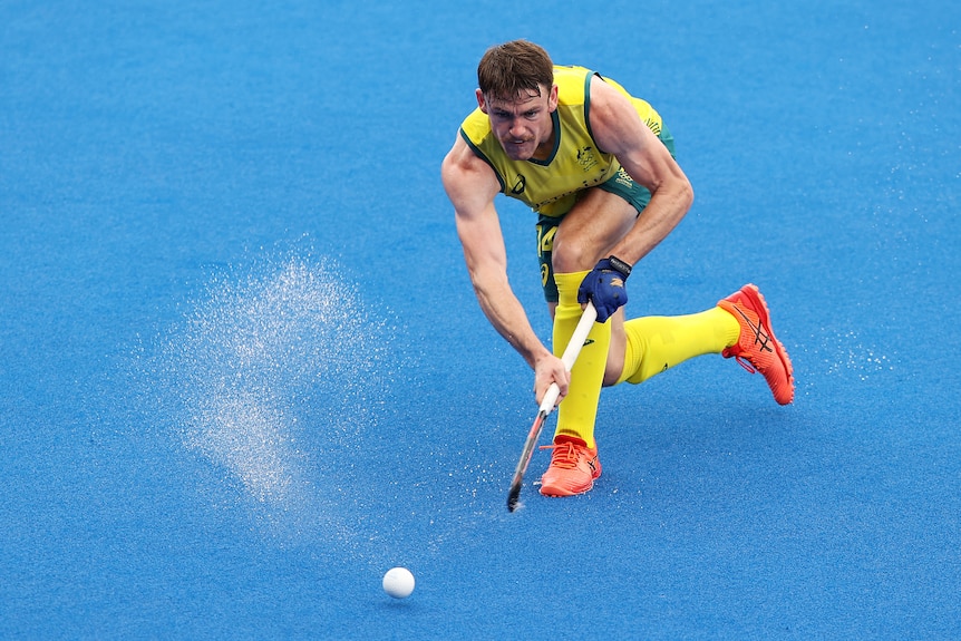 Man wearing yellow socks, green and yellow shorts and singlet, orange sneaker, chases a ball with a stick