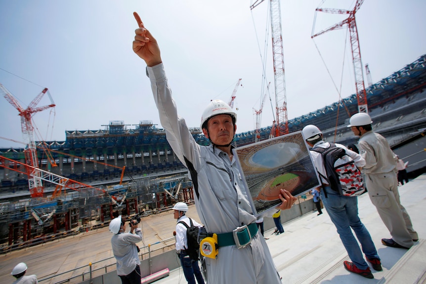 A Japanese man in a hard hat points at the sky in a half-constructed stadium 