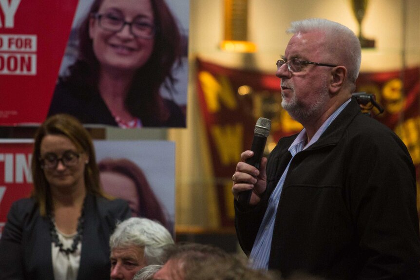 A man speaks into a microphone, standing in a seated crowd. Justine Keay is in the background in front of her campaign poster