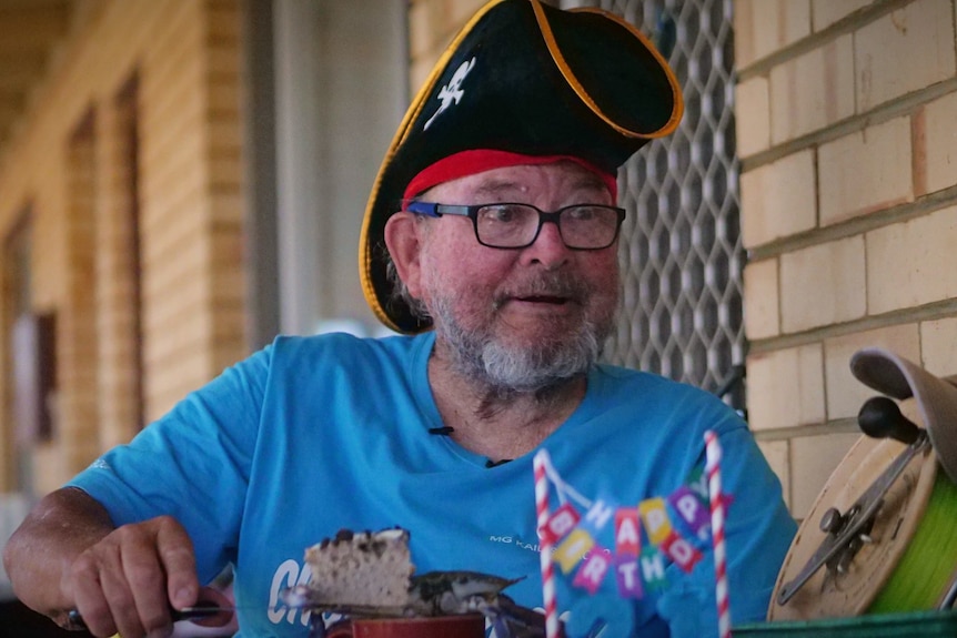 Elderly man, grey beard, glasses, in a pirate hat cutting a cake, smilings, looks off camera, wears blue tee.