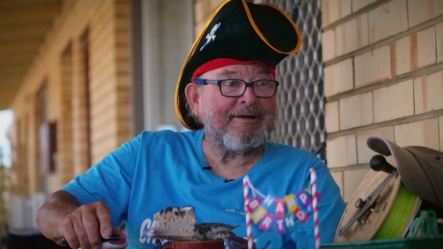Elderly man, grey beard, glasses, in a pirate hat cutting a cake, smilings, looks off camera, wears blue tee.