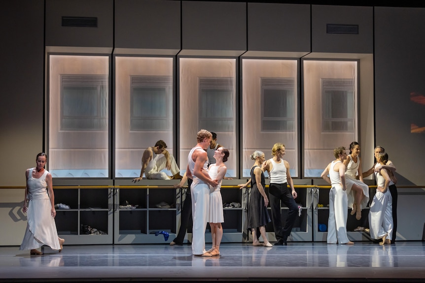 A group of dancers in white in a rehearsal room. A male and female dancer embrace in the centre of the shot.