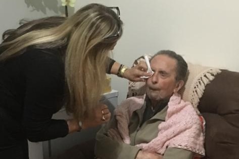 A woman with long blonde hair cleans the eye of an elderly seated man.